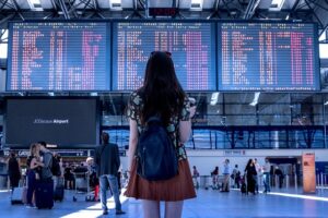 A person with a backpack stands facing away from the camera, looking up at large digital flight information display boards in a bustling airport terminal