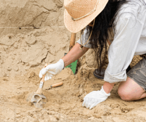 An archaeologist in a straw hat using a small brush to carefully uncover a human skull at a sandy excavation site.