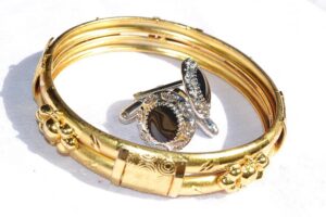 Two shiny gold bangles with intricate designs are displayed on a white surface, with a pair of ornate silver cufflinks with black centers resting inside them