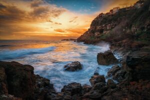 A dramatic coastal scene at sunset, with waves crashing against a rocky shore and cliffs