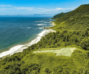Aerial view of a lush green coastline with waves crashing on a beach and a clearing with a small court