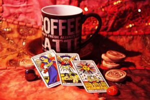 three colorful Tarot cards (The Temperance, The Judgment, and The Sun) laid out on a patterned red and gold fabric, with a black "COFFEE LATTE" mug