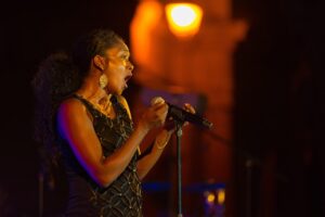 A female singer with dark, curly hair in a high ponytail, wearing a black, embellished dress and large hoop earrings, sings passionately into a microphone on a dimly lit stage