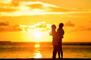 A silhouetted couple embraces on a beach at sunset, holding a glass of champagne