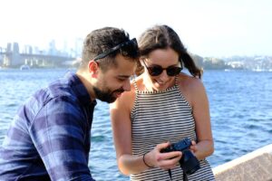 happy couple by a body of water with a city skyline in the background; the woman is showing the man something on a camera, and they are both smiling