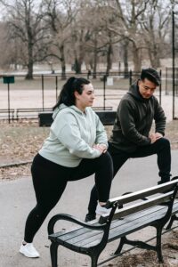 man and a woman in athletic wear are performing lunges outdoors in what appears to be a park