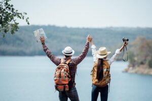 couple with raised arms overlooking a vast body of water surrounded by mountains, with one holding a map and the other a camera