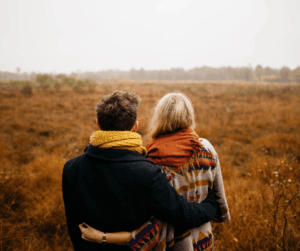 A couple, backs turned, hugging and looking out over a brown, grassy field on an overcast day