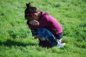 A young child in a purple hoodie is crouched down on a grassy field, with their head in their arms, appearing to be crying