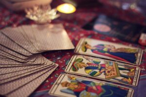 soft-focus shot of tarot cards, some face up revealing classic Marseille-style illustrations, and a stack of face-down cards, all resting on a patterned red fabric, with a blurred candle flame in the background