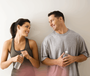 A man and a woman in athletic wear stand side-by-side against a plain white wall, holding water bottles and smiling warmly at one another.
