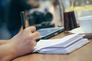 a person's hand writing in a notebook with a pen on a wooden table, with a camera, tablet, and drinks blurred in the background