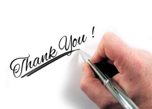 A close-up of a hand holding a pen, writing "Thank You!" in cursive script on a white surface