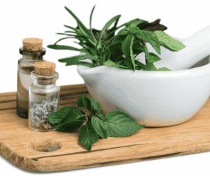 A small wooden cutting board holds a white mortar and pestle filled with fresh green herbs, and two small corked bottles containing dried herbs