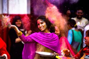 A joyful indian lady covered in vibrant pink and yellow powder, smiles broadly while celebrating Holi, with a cloud of colorful dust behind her