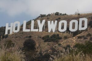 A wide shot of the iconic white "HOLLYWOOD" sign on a dry, grassy hillside under a partly cloudy sky