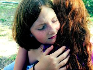 a young girl with freckles and closed eyes, embracing another person (whose back is visible), conveying a sense of comfort or emotion