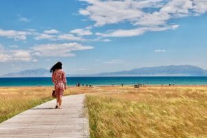 A person in a floral dress walks along a wooden boardwalk through tall golden grass, leading towards a large, clear blue lake