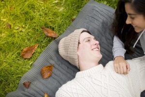 A man wearing a knit hat and sweater is lying on a gray blanket on green grass, looking up and smiling at a woman who is partially visible beside him