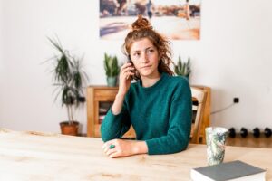 A young woman with reddish hair and a green sweater talks on a cellphone while sitting at a wooden table