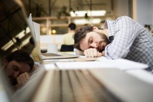 A man with a beard is sleeping with his head resting on a desk, next to a laptop, in an office environment