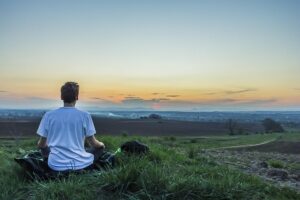 A man sits on a grassy hill, facing away from the camera, and meditates while watching the sunset