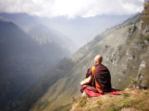 a bald monk, seen from behind, sitting on a red mat at the edge of a grassy cliff, gazing out at a vast, hazy mountain valley