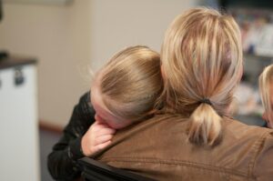 child with blonde hair is leaning their head on the shoulder of her mother