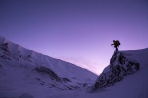 A solitary mountaineer with a backpack and trekking poles is silhouetted against a gradient purple sky, standing on a snowy cliff edge