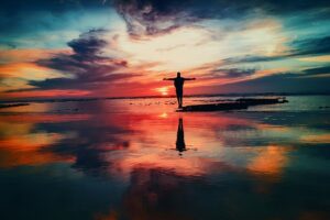 silhouette of a person standing on a reflective wet beach with arms outstretched, facing a dramatic sunset