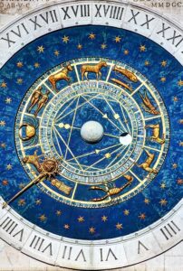 A detailed close-up of a large, elaborate astrological clock face, featuring a deep blue background with golden zodiac symbols, Roman numerals, and intricate gears.