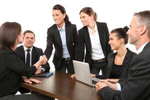 group of business professionals in a meeting setting, with two women in the foreground shaking hands and smiling