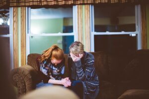 Two women sit on a couch, with their heads bowed in what appears to be sadness or prayer