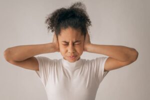 A young Black girl with curly hair, wearing a white t-shirt, covers her ears with her hands and scrunches up her face as if in discomfort from a loud noise