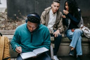 A young man wearing headphones is intently writing in a notebook outdoors on a bench, while two other individuals are seated and talking at his back