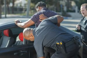 Police officer conducting a search or arrest, pressing person against a car while another officer stands nearby.