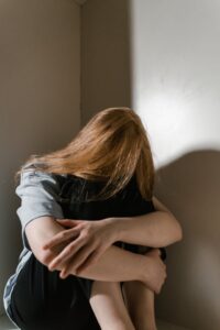 A person with long, reddish hair is curled up in the corner of a room, with their face hidden and their arms wrapped around their knees, suggesting sadness or distress