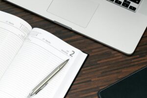 An open planner with a silver pen resting on it, placed on a wooden desk alongside a laptop and a black leather notebook