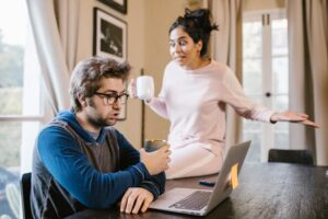 A man wearing glasses sits at a table with a laptop looking surprised, while a woman in pajamas sits on the table behind him, gesturing with an expression of exasperation