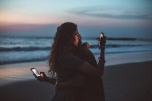 two individuals embracing on a beach at dusk, both engrossed in their smartphones during an embrace