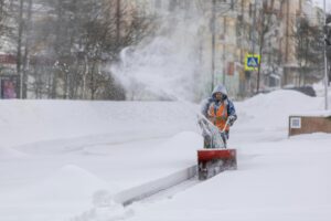 A worker in a hooded jacket and orange safety vest clears snow from a sidewalk using a snow blower on a cold, snowy day in the city.