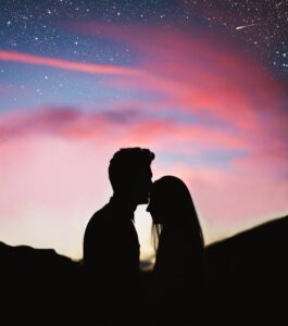  silhouette of a couple, with the man kissing the woman's forehead, against a vibrant sunset sky filled with stars