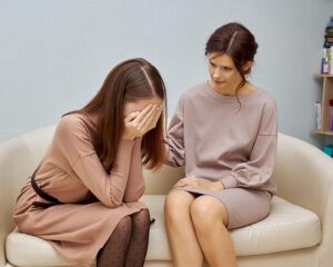 two women on a light-colored sofa. One woman is sitting with her face buried in her hands, appearing distressed and crying. The other woman, is sitting next to her, looking at her with a concerned expression