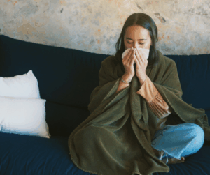 A person wrapped in a green blanket sitting on a sofa and sneezing into a tissue.