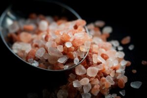 pink Himalayan salt crystals, mostly a mix of light pink, orange, and white, spilled from a metal measuring spoo