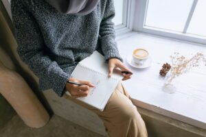 a person from a high angle, dressed in a grey sweater and a hijab, sitting by a window ledge and writing something in a notebook
