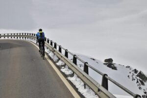 person on a bicycle rides along a winding asphalt road with a metal barrier, surrounded by vast snow-covered slopes under an overcast sky