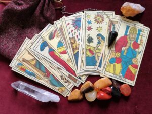 tarot cards spread out on a burgundy cloth alongside a dark pendulum, a clear quartz crystal, and an assortment of polished colorful gemstones
