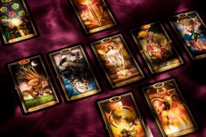 A collection of ornate tarot cards, some of which are identifiable as "Ten of Wands," "Death," "Justice," "King of Cups," "The Hanged Man," "Five of Pentacles," "Ace of Wands," and "The Devil," laid out on a deep purple, textured fabric