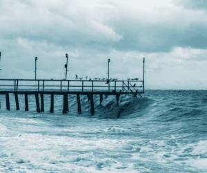 An image of a large wave crashing against the wooden support beams of a pier under a cloudy sky